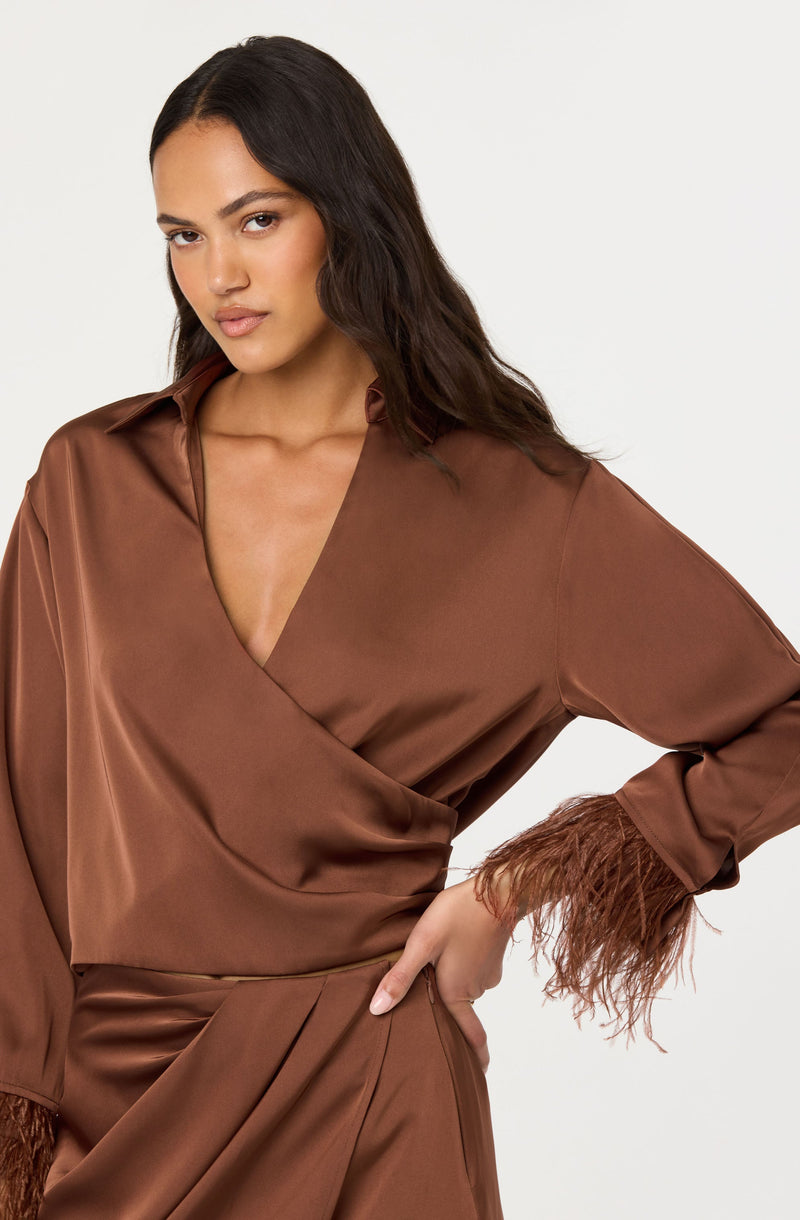 The woman is wearing a satin brown top with a V-neckline and a wrap design. The top features long sleeves with feathered cuffs at the ends. She is also wearing a matching satin brown skirt with pleats at the waist.