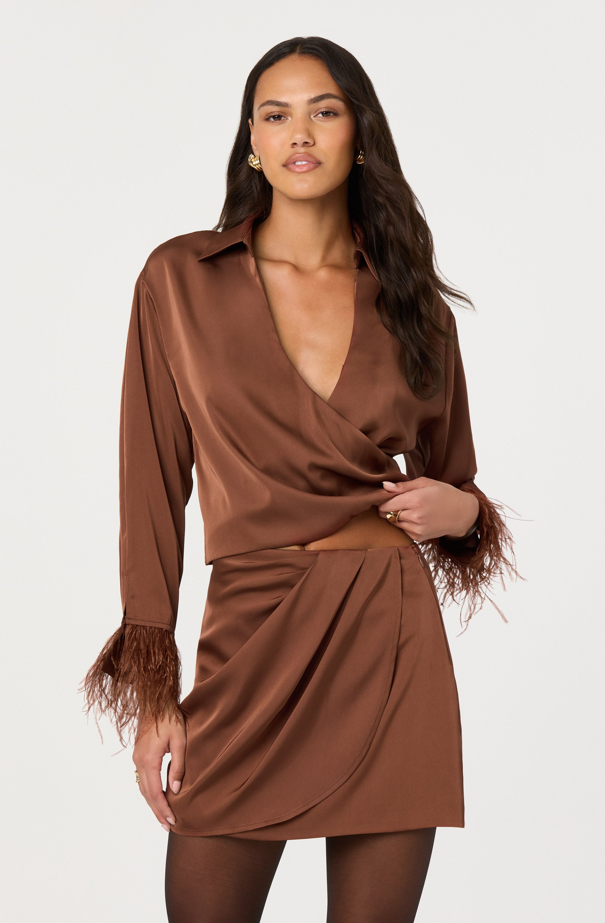 A woman is wearing a silky, satin-like brown blouse with a V-neck and long sleeves that have feathered cuffs. She is also dressed in a matching brown skirt with pleats, a draped detail at the front, and a feathered trim at the hem.