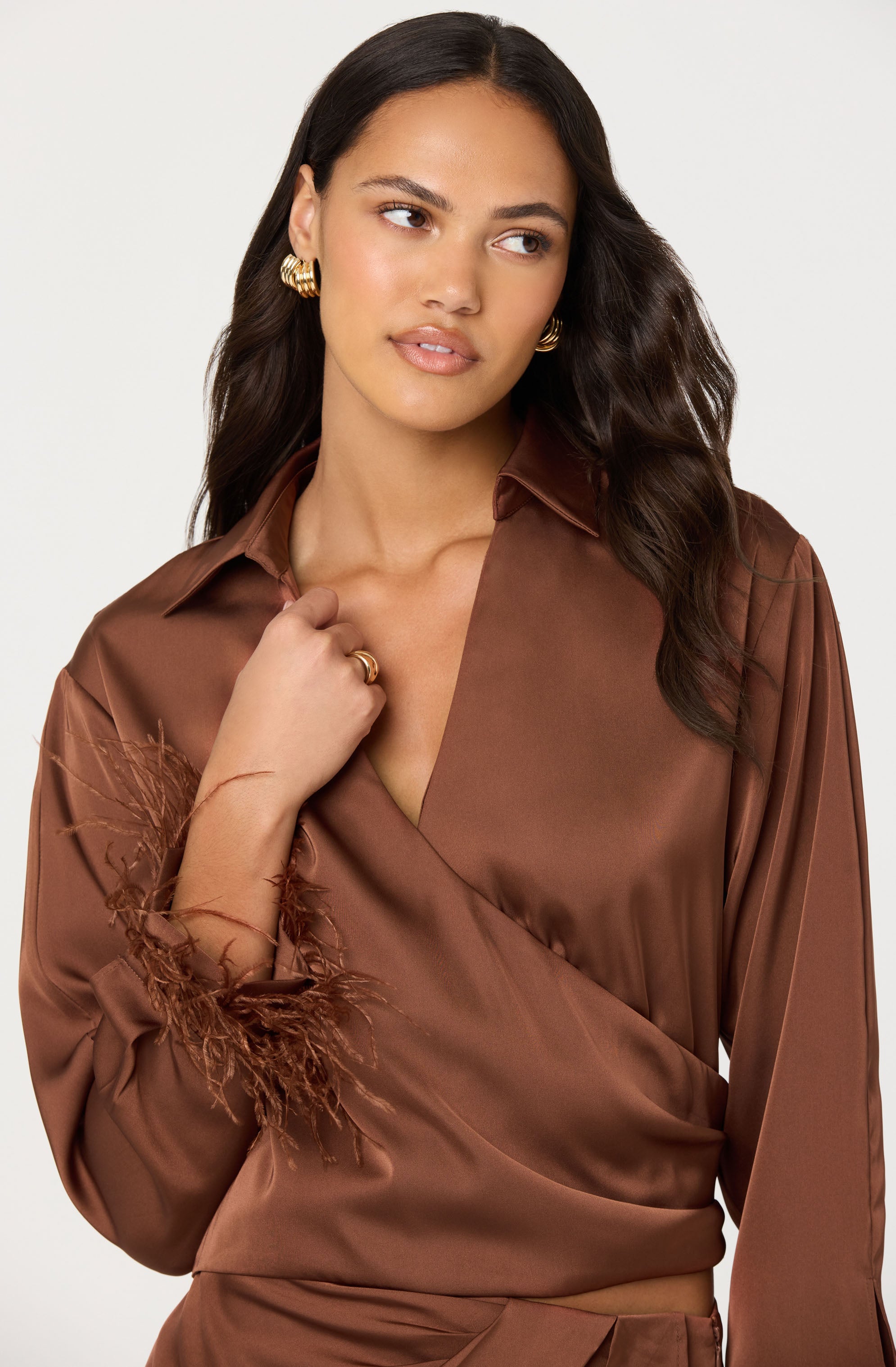 A woman is wearing a silky, brown wrap blouse with a collar. The blouse has decorative feathered details at the cuffs of the sleeves.