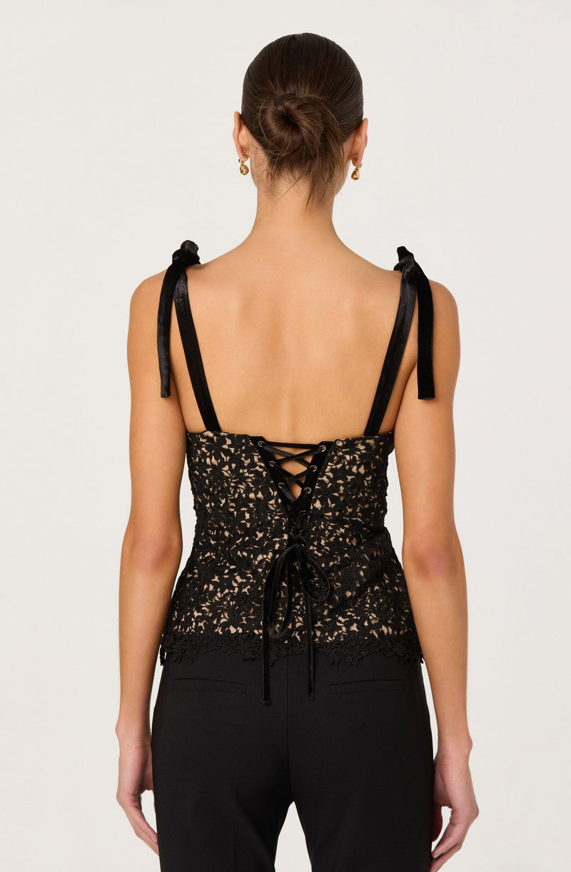 A woman is wearing a sleeveless black lace top with intricate floral patterns. The top features black straps tied into bows on her shoulders and a lace-up detail at the back with black laces crisscrossing and tied at the bottom. She is also wearing black pants. Her hair is styled in a neat bun, and she has gold earrings.
