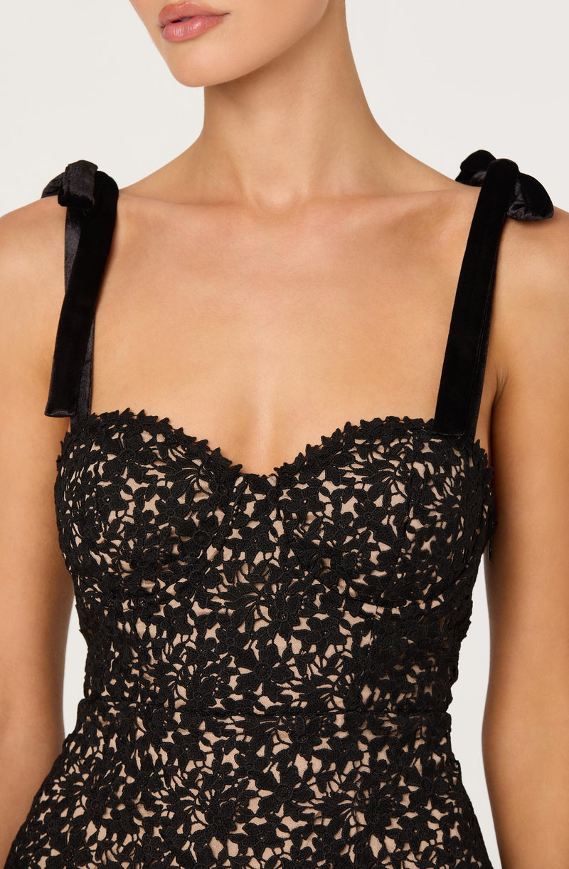 A woman is wearing a sleeveless dress with black lace overlay featuring a floral pattern. The dress has black satin shoulder straps tied into bows at the shoulders. The bodice is fitted, showcasing the intricate lace design against a nude-colored lining.