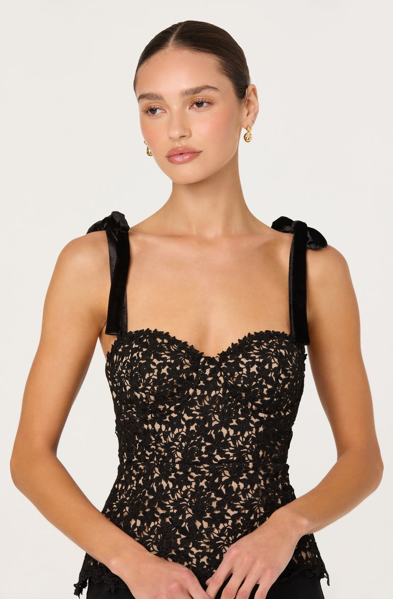 A woman is wearing a sleeveless black lace top with intricate floral patterns. The top features shoulder straps made of black ribbon tied into bows.