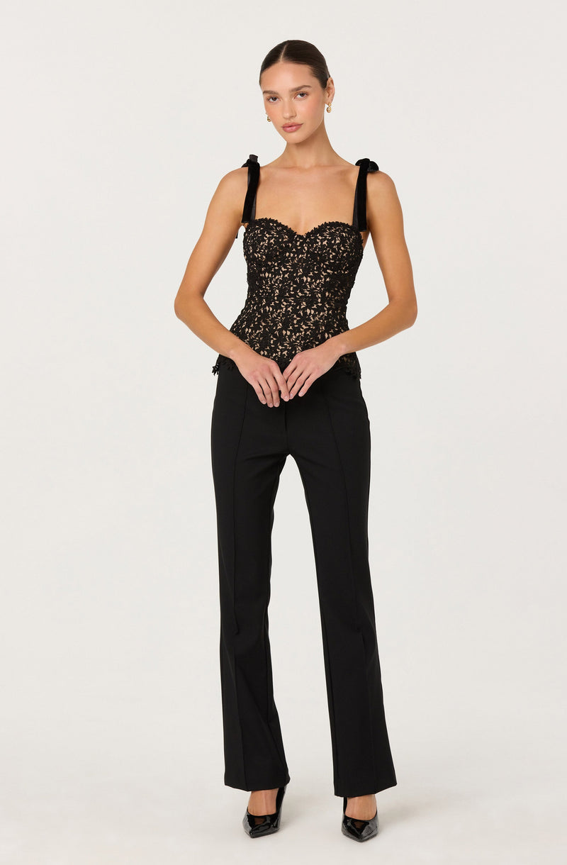 The woman is wearing a black lace top with scalloped edges along the neckline and straps tied into bows at the shoulders. She pairs it with black fitted dress pants that have a straight-leg cut, and is wearing black high-heeled shoes.
