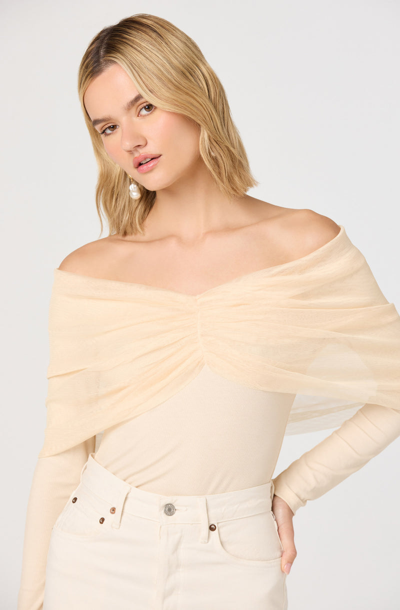 A woman is wearing an off-the-shoulder top made of a light, sheer fabric in a soft cream color. The top has a gathered detail at the neckline, creating a draped effect that wraps around her shoulders and upper arms. She pairs it with high-waisted cream-colored pants that have a button and zipper closure, and visible front pockets.