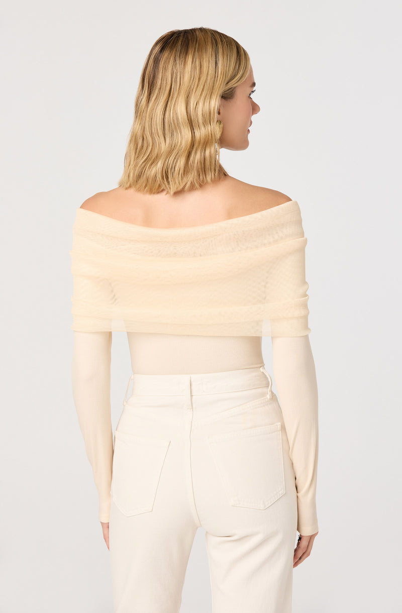 A woman is wearing an off-the-shoulder, light-colored top with a textured, sheer overlay that drapes around her shoulders. She is also wearing high-waisted, cream-colored pants with visible back pockets.