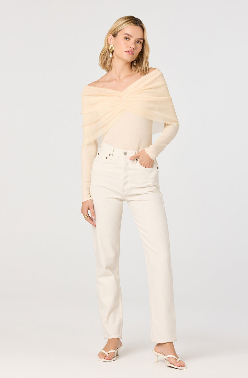 A woman is wearing a light beige, off-the-shoulder top made of a soft, sheer fabric that is draped around her shoulders. She has paired it with high-waisted, straight-leg white jeans. Her footwear consists of white, open-toe high-heeled sandals. She accessorizes with large gold hoop earrings and a ring on her right hand.