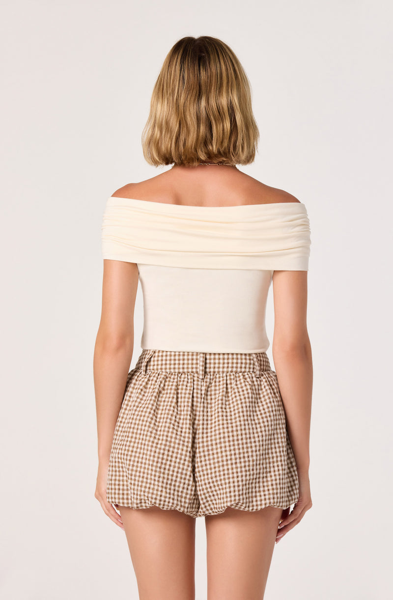 Woman wearing an off-the-shoulder cream top and beige checkered shorts.