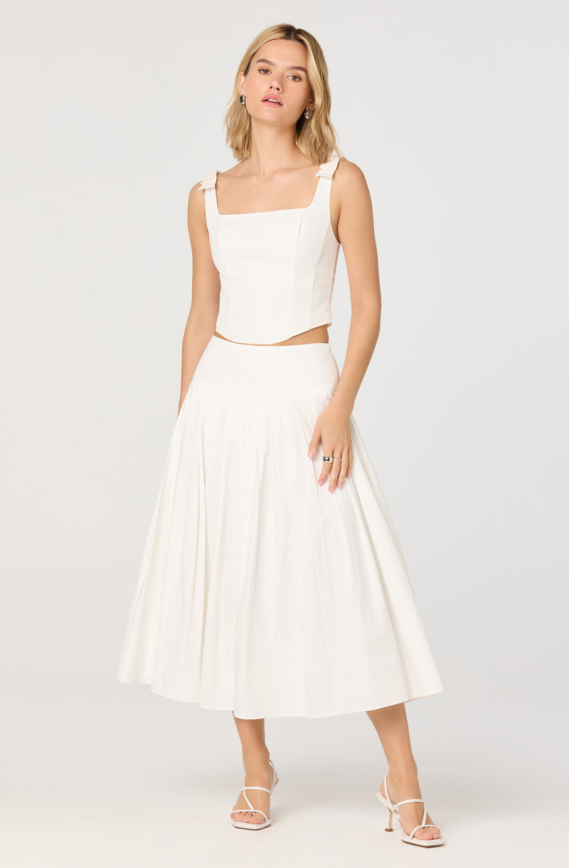 A woman is wearing a sleeveless, white crop top with wide shoulder straps tied into bows. She pairs it with a high-waisted, white, pleated midi skirt that reaches mid-calf. She is also wearing white open-toe high-heeled sandals.