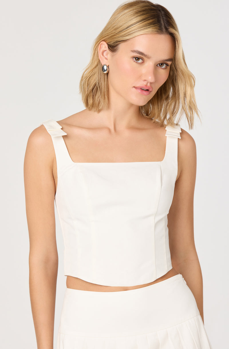 A woman is wearing a sleeveless, white crop top with wide shoulder straps that feature small bow details. The top has a straight neckline and is fitted, ending just above the waist. She is also wearing a matching white skirt.