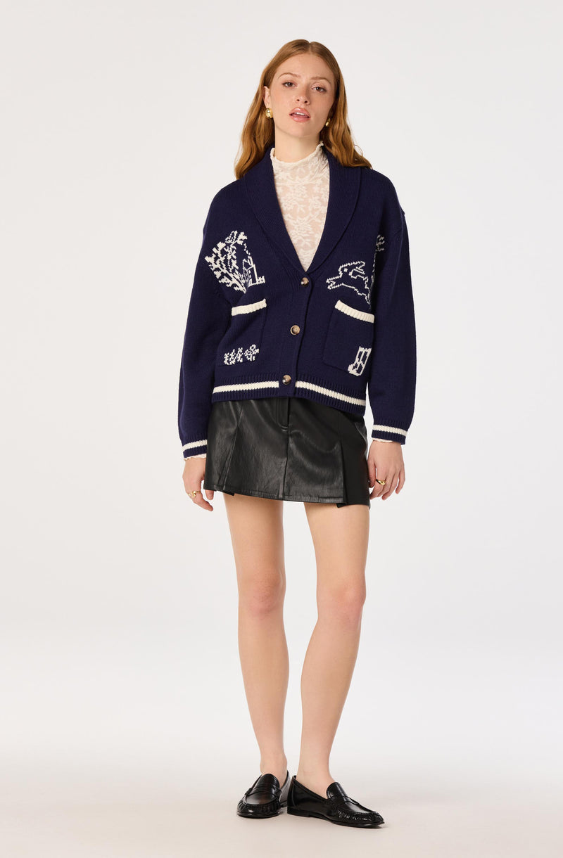 Woman wearing a navy blue cardigan with white patterns, a sheer lace top, black leather skirt, and black loafers.