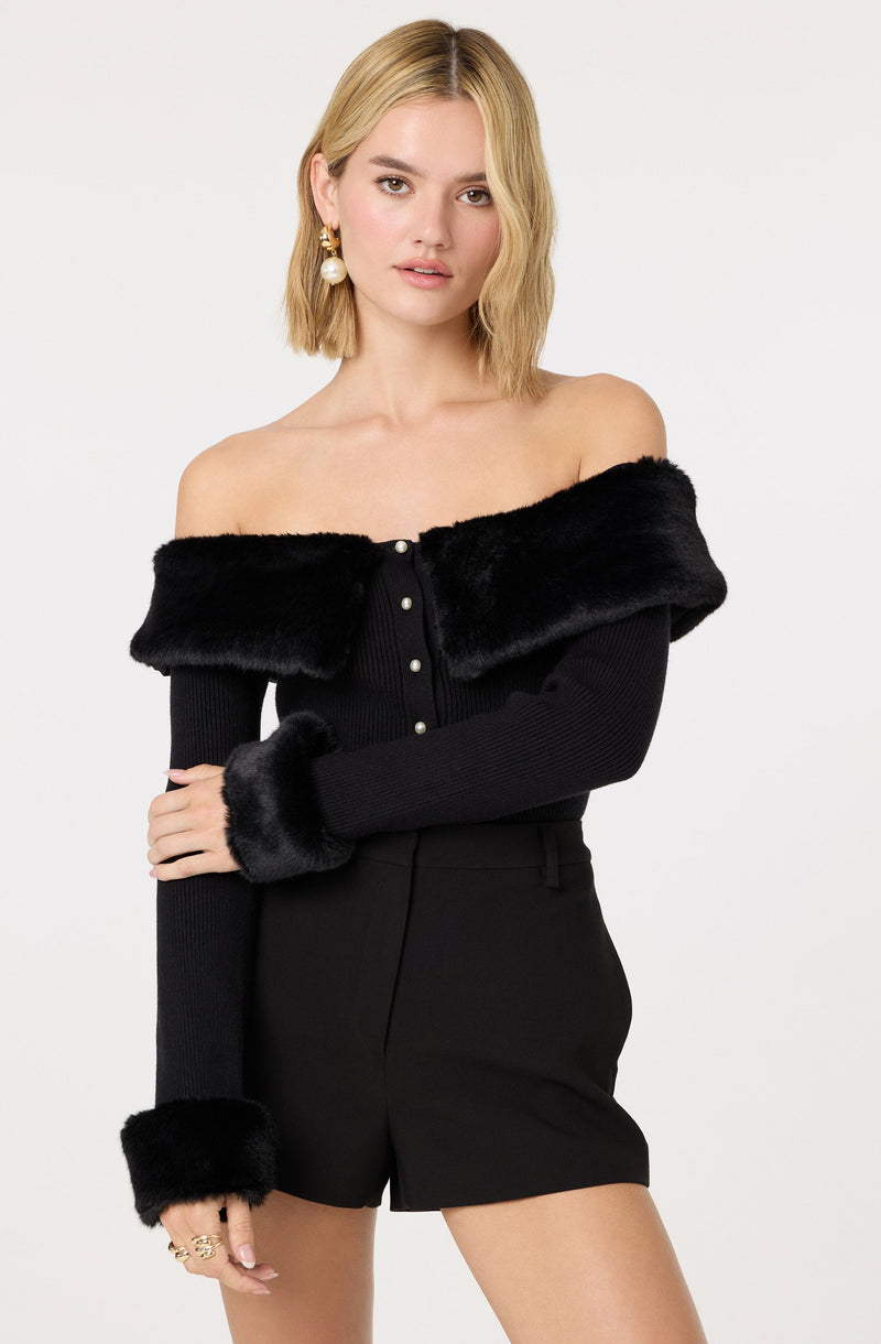 A woman is wearing a black off-the-shoulder sweater with a wide, furry collar and matching furry cuffs on the sleeves. She is also dressed in high-waisted black shorts.