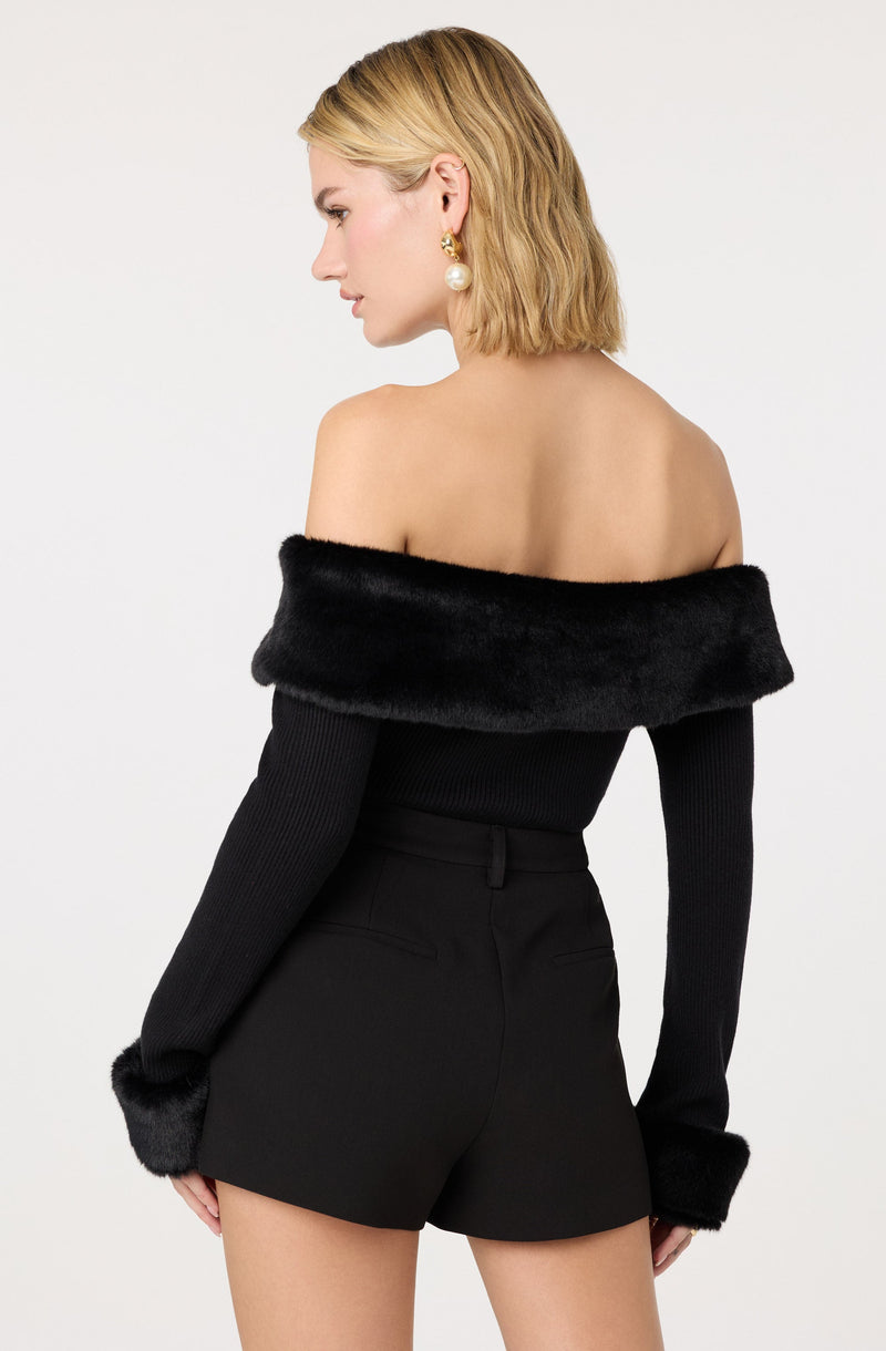 A woman is wearing an off-the-shoulder black sweater with a wide, plush black fur collar that drapes over her shoulders. The sweater has long sleeves with matching black fur cuffs. She pairs the sweater with high-waisted black shorts made of a fitted fabric. She accessorizes with gold earrings featuring large pearl drops and has shoulder-length blonde hair styled in loose waves.