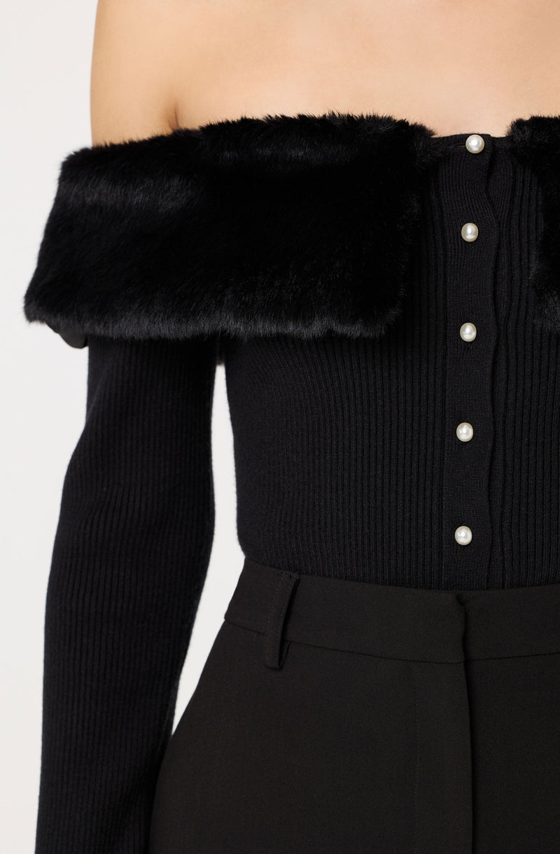 A person wearing a black off-the-shoulder top with a wide, faux fur collar. The top features a row of decorative pearl buttons down the front. They are also wearing high-waisted black pants.