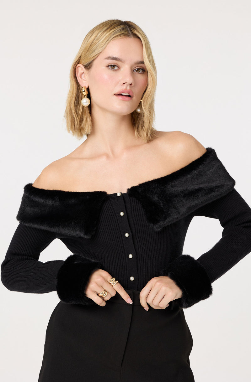 A woman is wearing a black off-the-shoulder top with faux fur trim along the neckline and cuffs. The top has a row of pearl-like buttons down the front. She accessorizes with large pearl drop earrings and multiple rings.