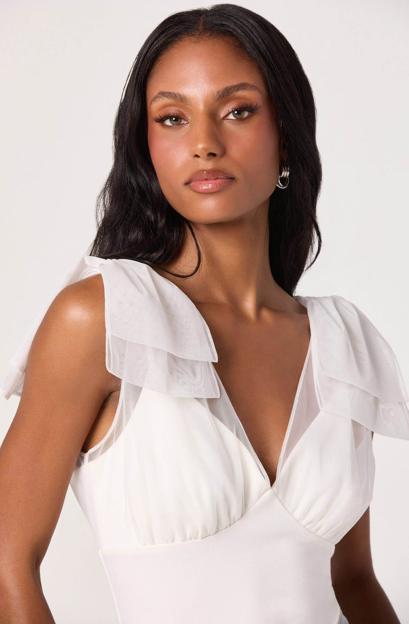 A woman is wearing a white sleeveless dress with ruffled, sheer fabric details on the shoulders. The dress has a v-neckline and is fitted at the waist.