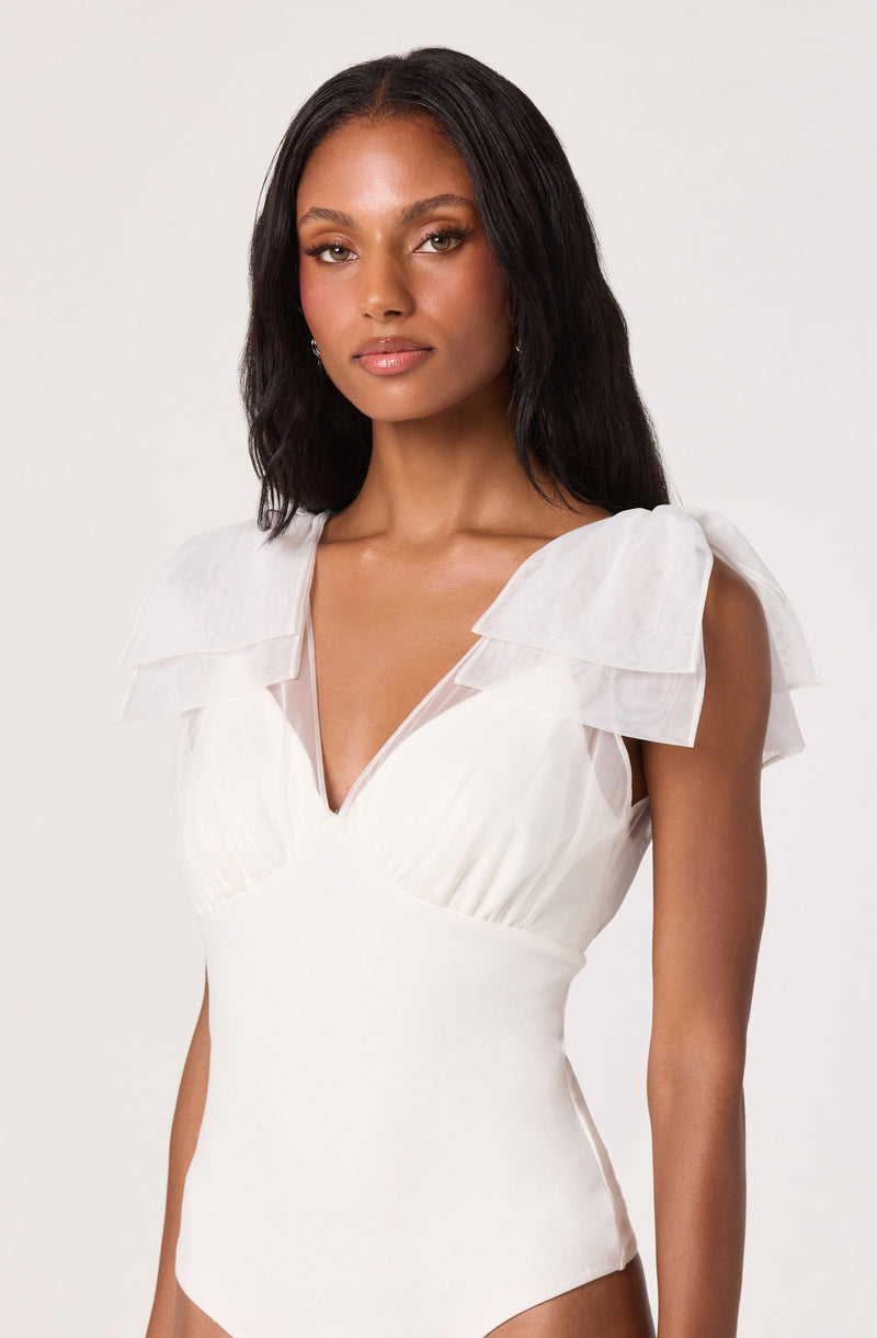 A woman is wearing a sleeveless, white bodysuit with a deep V neckline. The bodysuit features layered ruffle details on the shoulders and a gathered detail at the bust. The fabric appears smooth and fitted, emphasizing her silhouette.