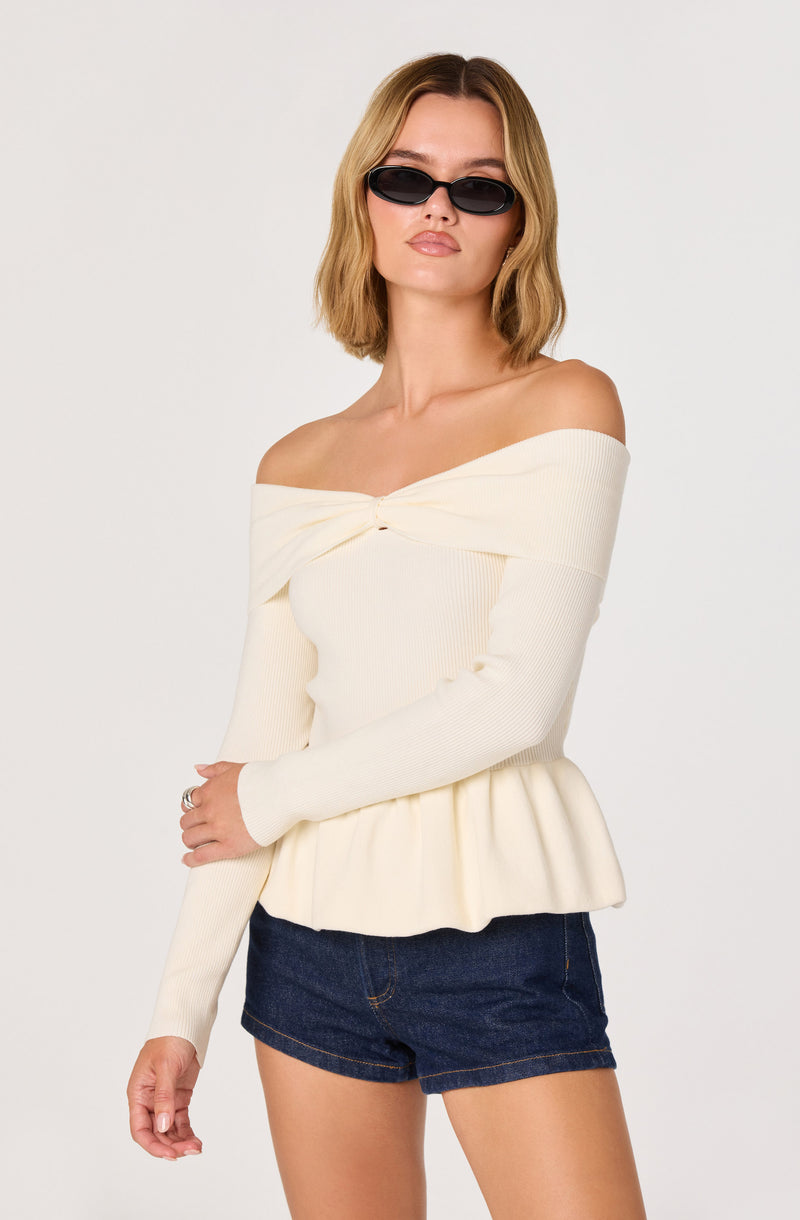 Woman wears off-the-shoulder cream top with a twisted knot detail and ruffled hem, paired with dark denim shorts.
