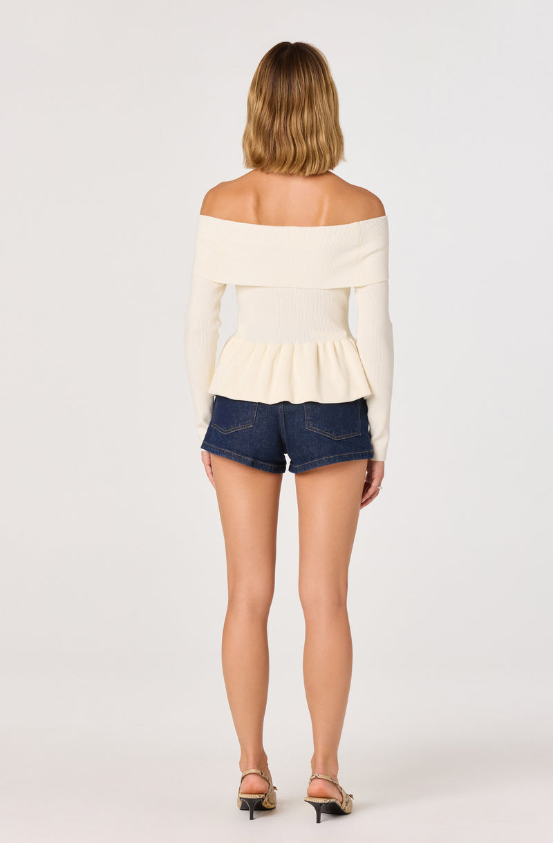 Off-the-shoulder white sweater with a ruffled hem, paired with dark denim shorts and beige heels.