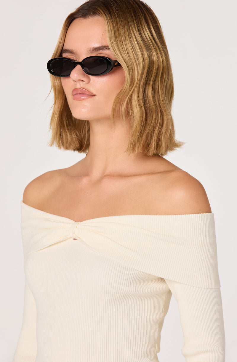 Woman in off-the-shoulder cream ribbed top and black oval sunglasses.