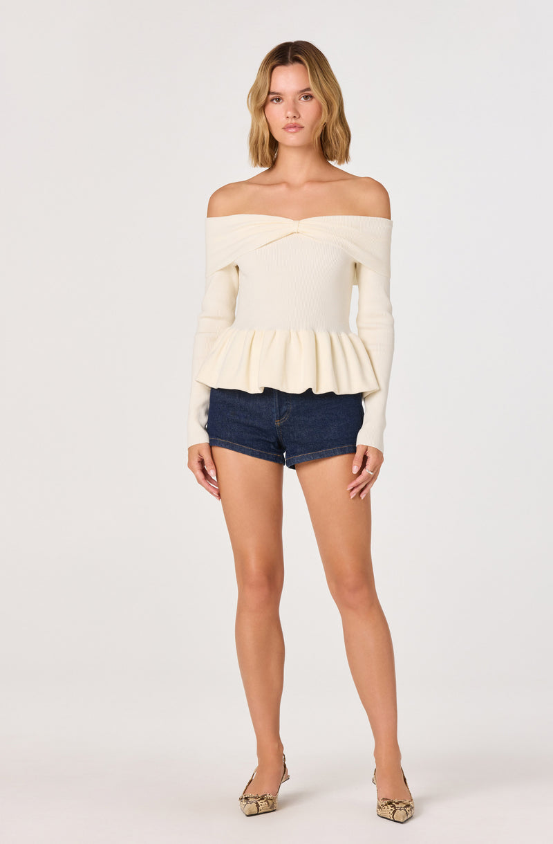 Off-shoulder cream sweater with ruffle hem, paired with dark denim shorts and snakeskin heels.
