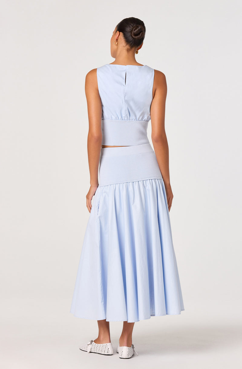 Woman in light blue sleeveless top and long full skirt, wearing white perforated shoes.