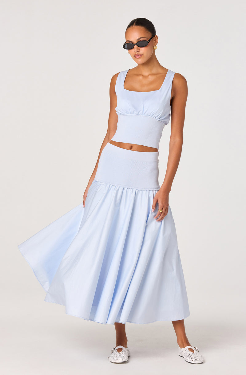 Woman in a light blue sleeveless crop top, matching high-waisted skirt, black sunglasses, white shoes.