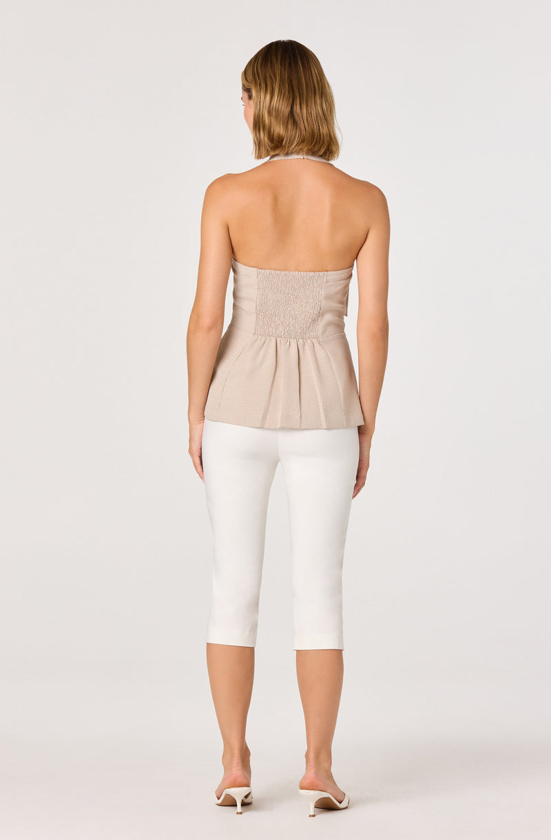 Back view of a woman wearing a strapless beige top with pleats and smocked waist, paired with white cropped pants and heels.