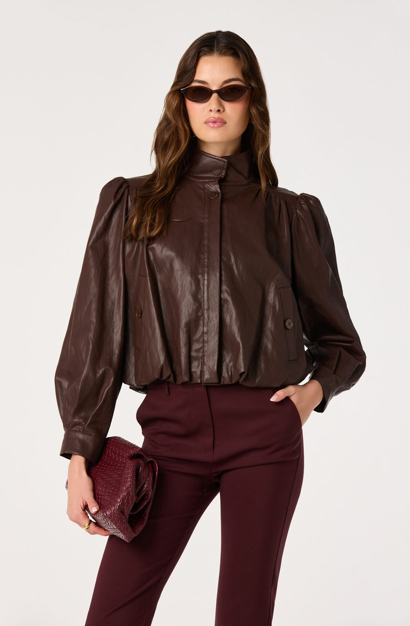 Woman wearing a brown leather jacket with puffed sleeves and high neck, burgundy pants, and holding a burgundy clutch.