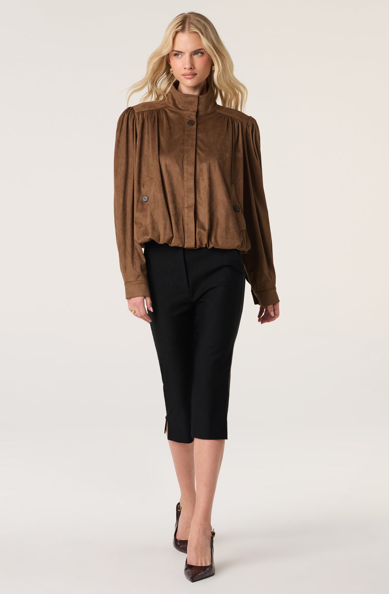 Woman wearing a brown, long-sleeved, high-neck jacket with pleats on shoulders and black pants.