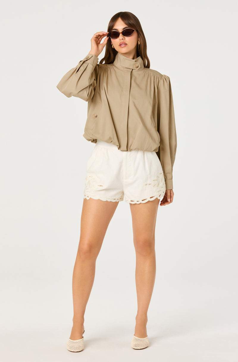 Woman wearing a beige high-neck blouse with puffed long sleeves and white eyelet shorts.