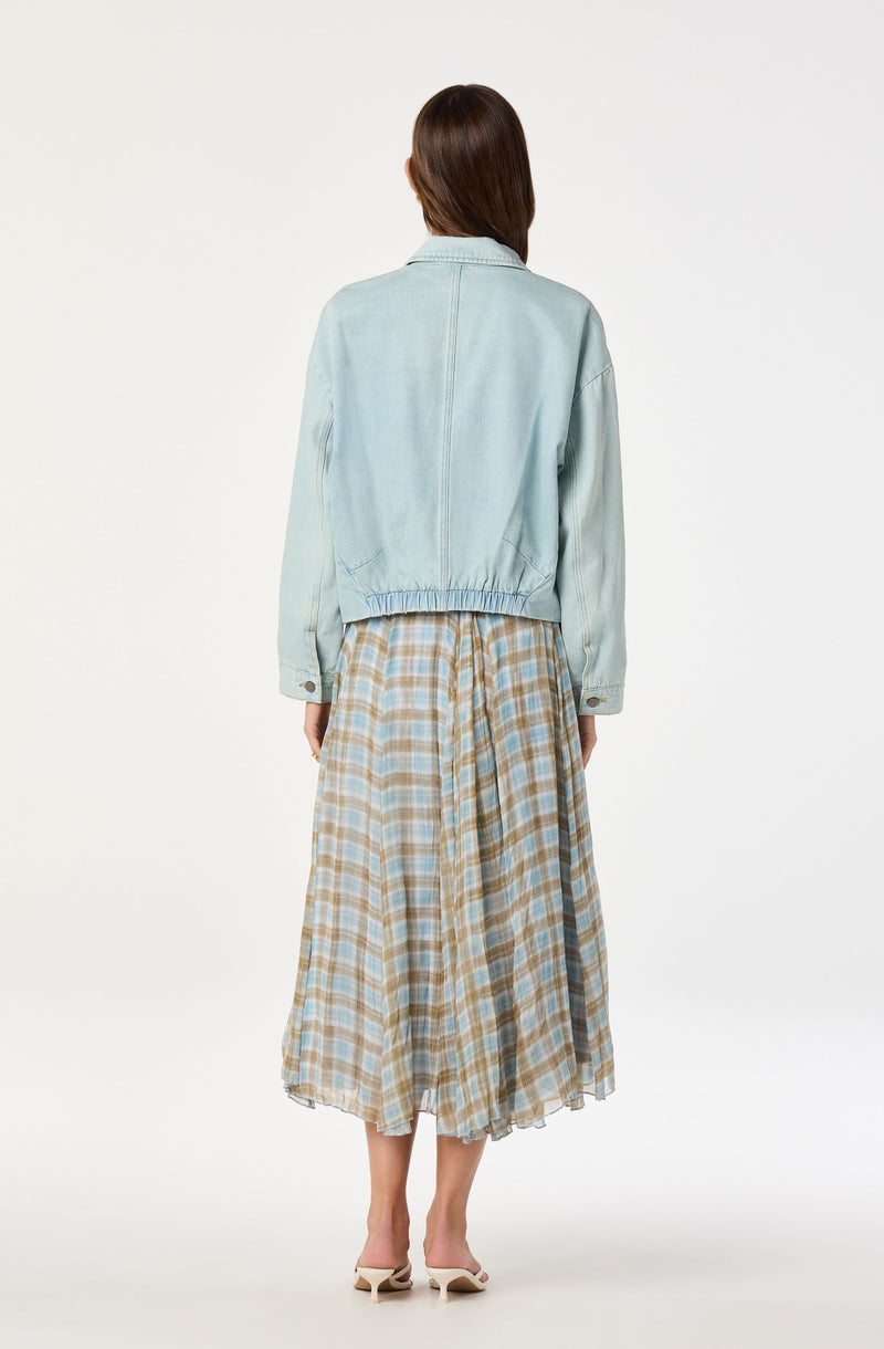 Woman wearing a light blue denim jacket and a beige plaid midi skirt with white heels.