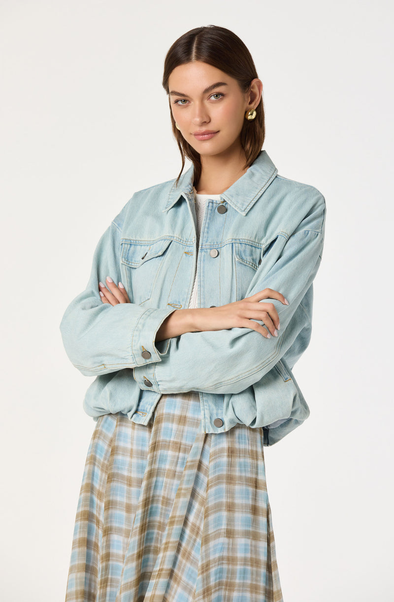 Young woman wearing a light blue oversized denim jacket and a plaid skirt with beige, blue, and white tones.