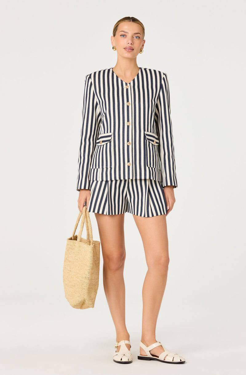 Woman in a navy and white vertically striped blazer and matching shorts, holding a straw tote bag.