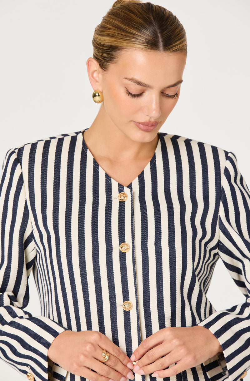Woman wearing a navy and white vertically striped blazer with gold buttons and gold earrings.