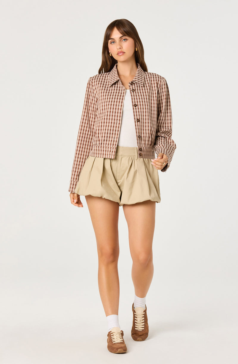 Woman wearing a checkered jacket, beige pleated shorts, white top, white socks, and brown sneakers.