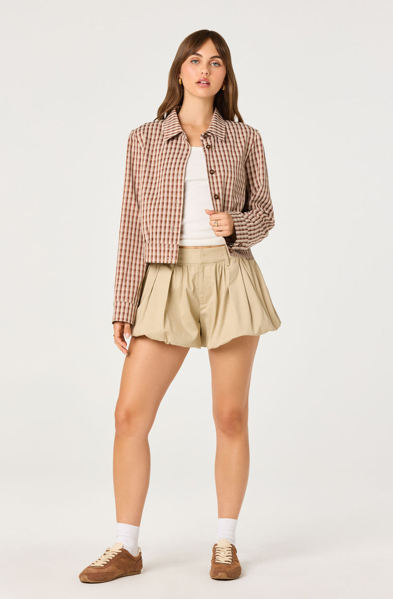 Woman wearing a checkered plaid jacket in shades of brown and beige, beige pleated shorts, white top, white socks, and brown sneakers.