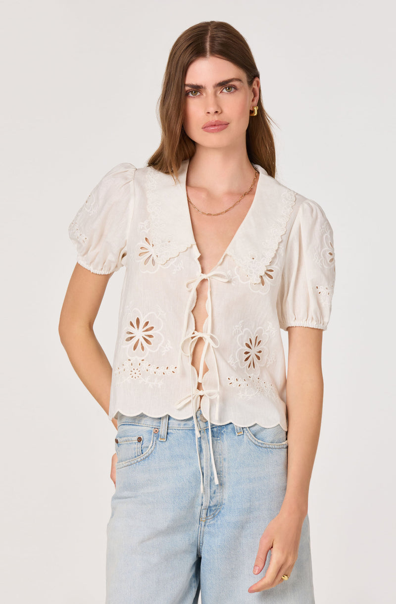 Woman wearing a white embroidered blouse with puff sleeves and scalloped hem, paired with light blue jeans.