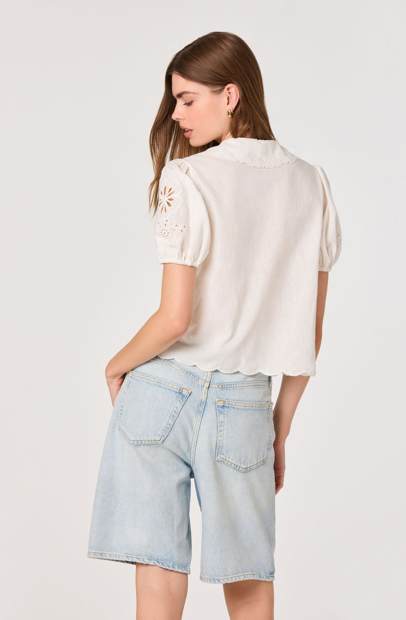 White blouse with eyelet embroidery on sleeves and scalloped hem, paired with light-wash denim shorts.