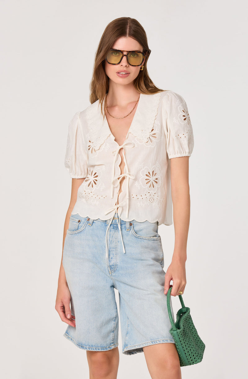 Woman in white embroidered blouse with scalloped edges, light denim shorts, and large sunglasses.