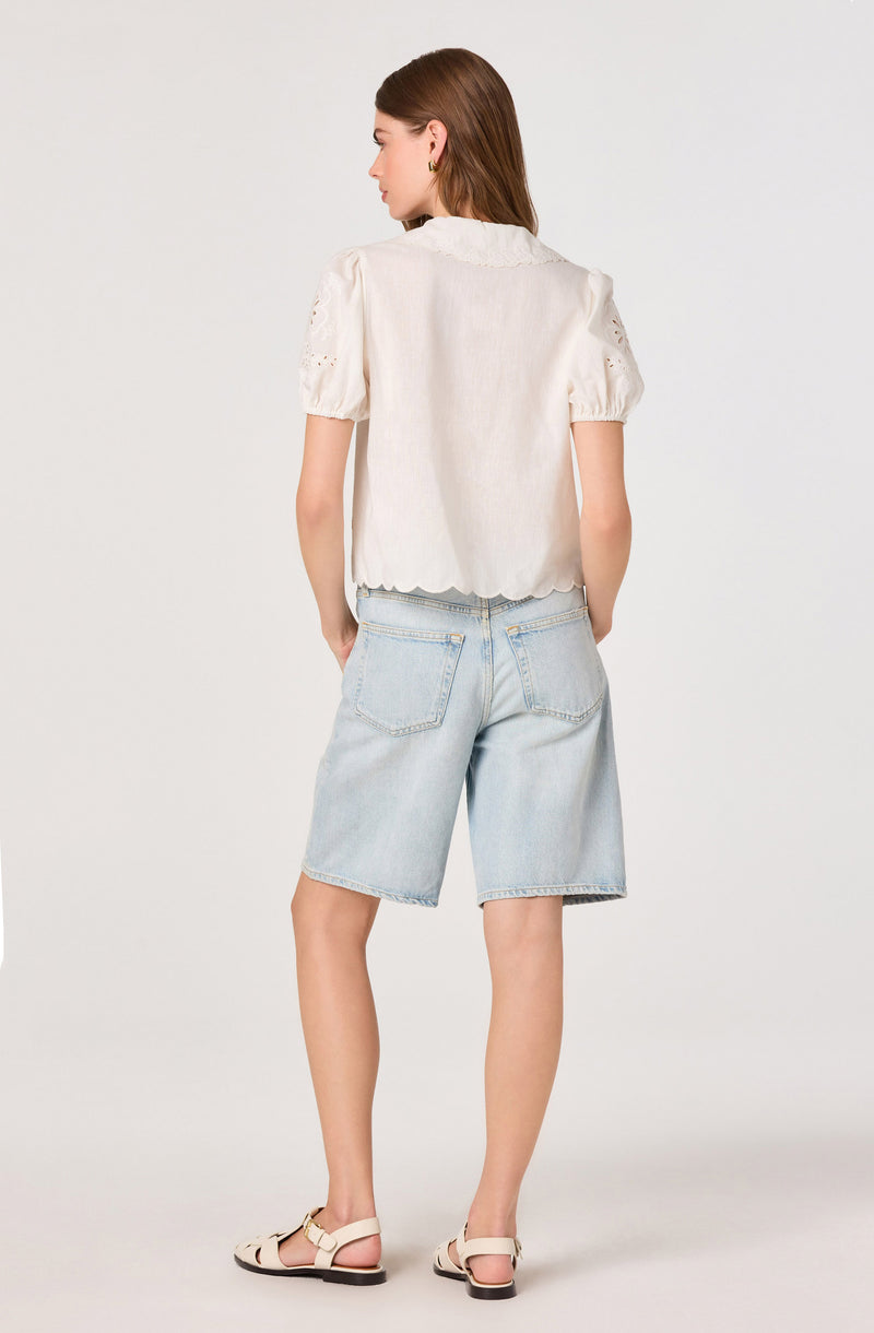 Woman wearing a white embroidered blouse with puff sleeves and scalloped hem, paired with light denim shorts and beige sandals.