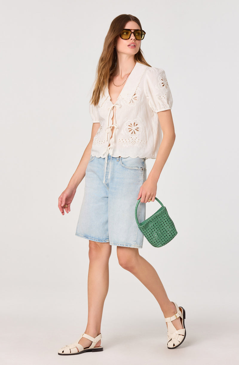 Woman wearing a white embroidered short-sleeve blouse, light denim shorts, beige sandals, yellow-tinted sunglasses, and holding a green woven handbag.