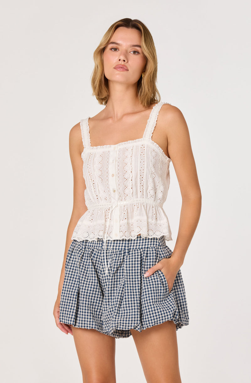 Woman in a white lace sleeveless top with ruffled hem and navy gingham shorts.