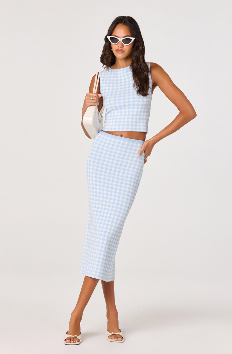 Woman in a blue and white gingham crop top and matching skirt, white sunglasses, holding a white handbag.