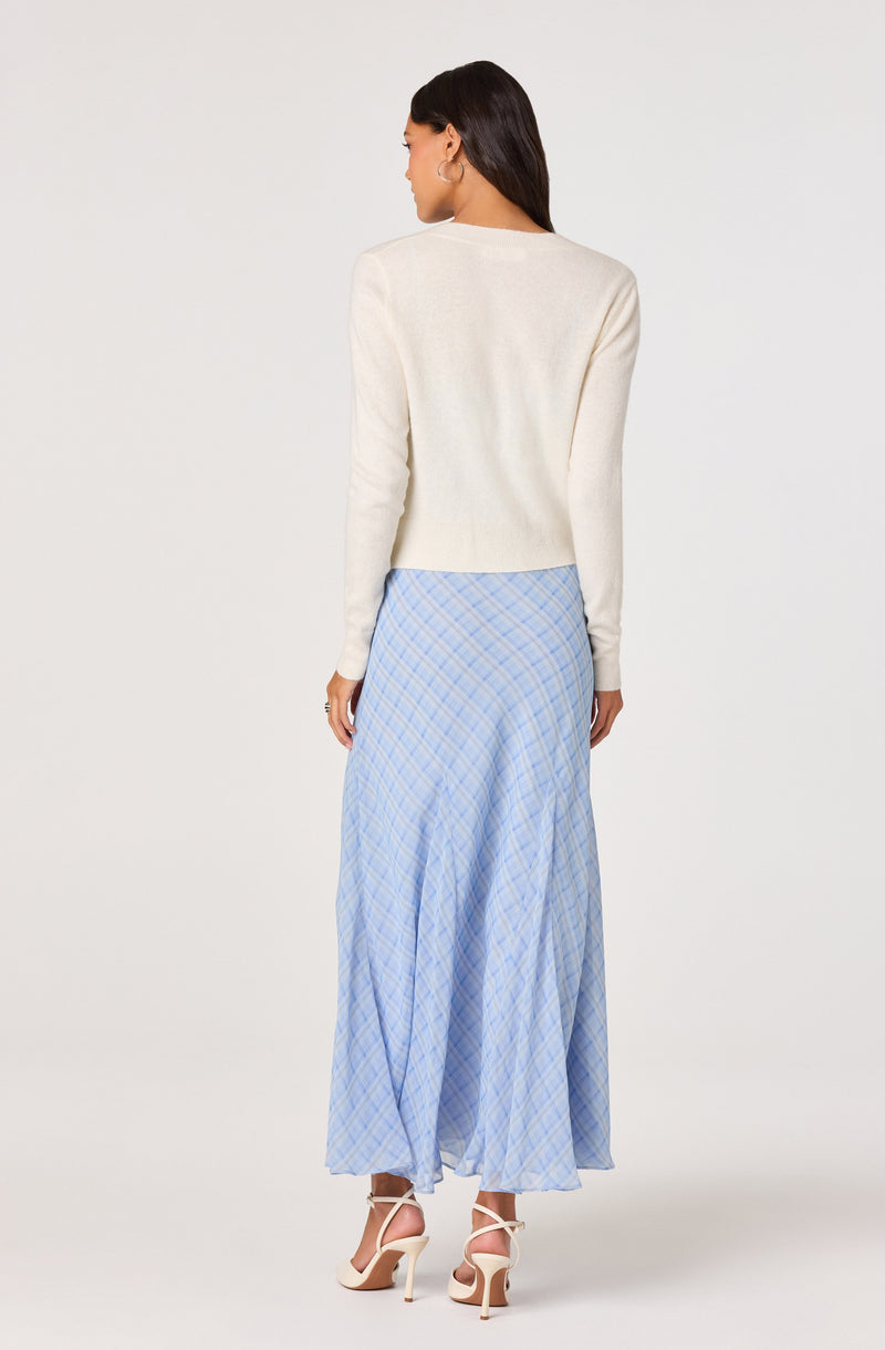 Woman wearing a white knit sweater and a long, plaid blue and white skirt, with white high-heeled sandals.