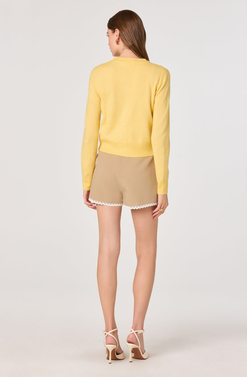 Woman in yellow sweater, beige shorts with white trim, and nude heels.