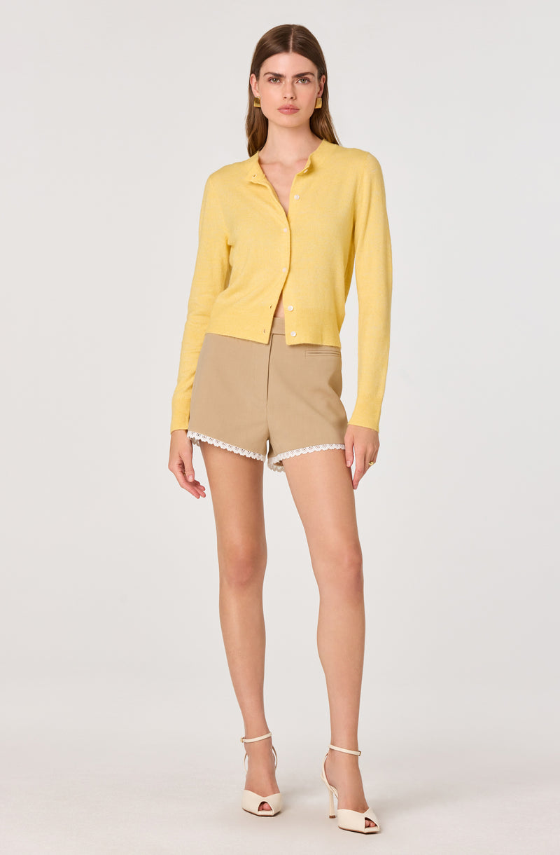 Woman in yellow cardigan, beige shorts with white lace, and white heels.