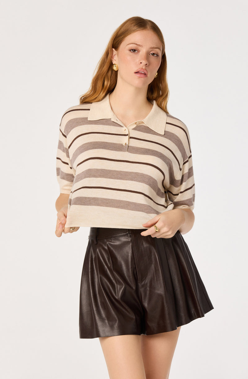 Woman wearing a beige and brown striped polo shirt and black leather pleated shorts.