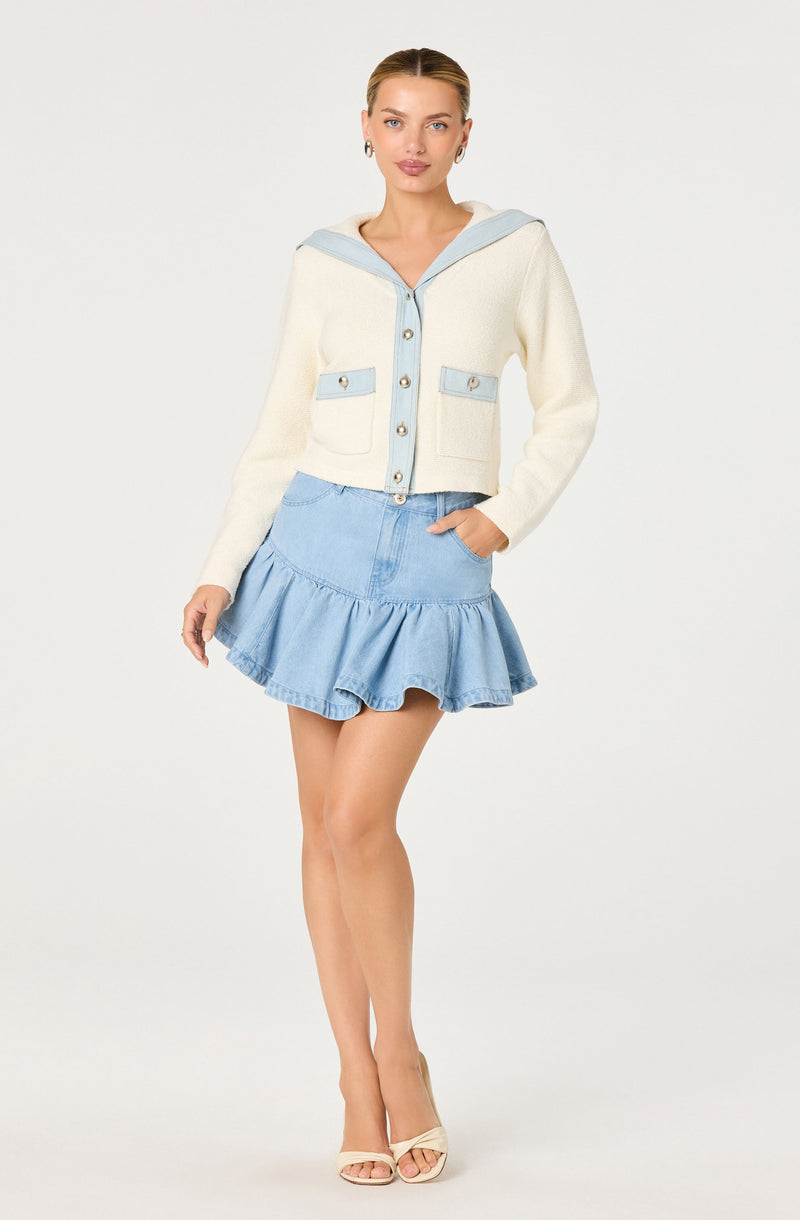 Woman in cream and light blue sweater with pockets and sailor collar, wearing a light blue ruffled denim skirt and beige high heels.