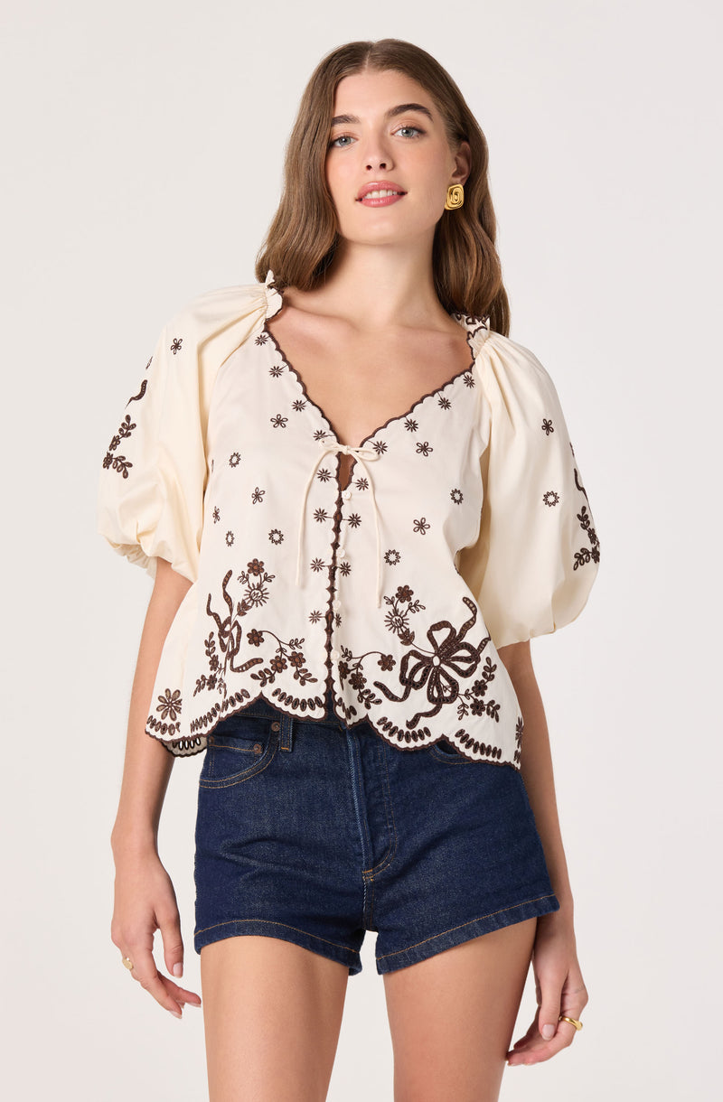 Cream blouse with puffy sleeves and dark floral embroidery, paired with dark denim shorts.