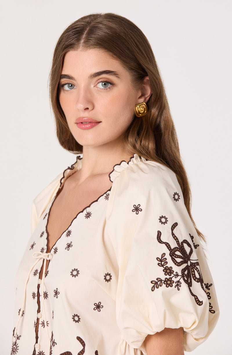 Woman wearing a cream-colored blouse with dark embroidered floral and bow designs, puffed sleeves.
